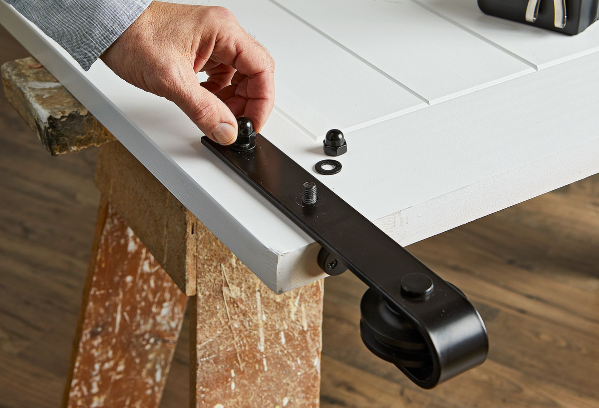A person's hand carefully attaching the black roller hardware to the top of a white barn door, securing it with a nut as part of the installation process.